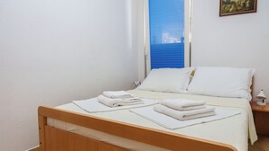 Een enkel bed met witte lakens en een houten frame, een nachtkastje met een lamp, en een raam met blauwe jaloezieën.