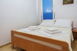 Un lit simple avec du linge de lit blanc et un cadre en bois, une table de chevet avec une lampe, et une fenêtre avec des stores bleus.