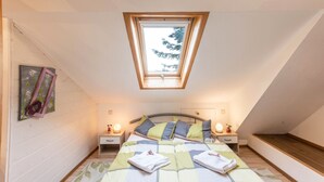 Ein Schlafzimmer mit schräg verlaufender Decke, einem Oberlicht, einem Bett mit Kissen, Nachttischen mit Lampen und einer hölzernen Bank.