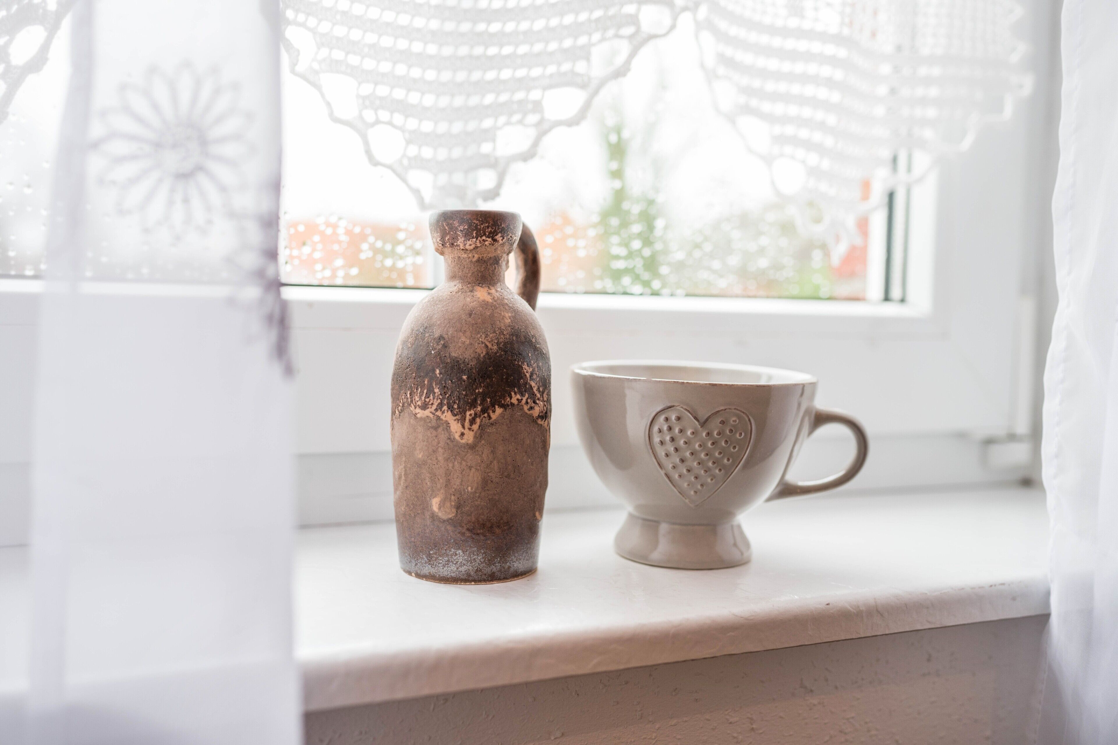 Une cruche en céramique rustique et une tasse en forme de cœur sont posées sur un plan de travail blanc, près d’une fenêtre ornée de rideaux en dentelle.