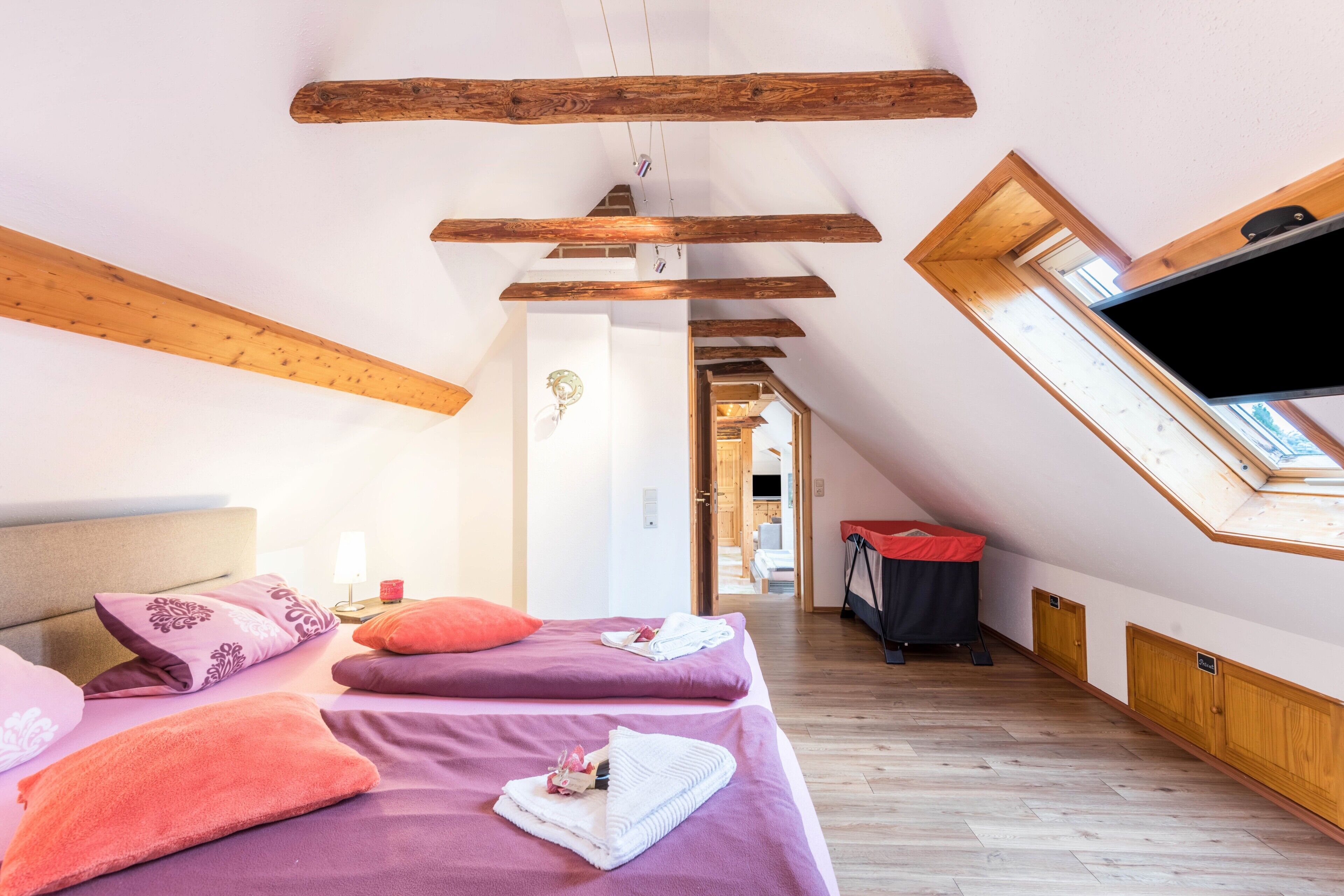 Una habitación con techo inclinado, vigas de madera, una cama con ropa de cama púrpura, un televisor de pantalla plana y un tragaluz.