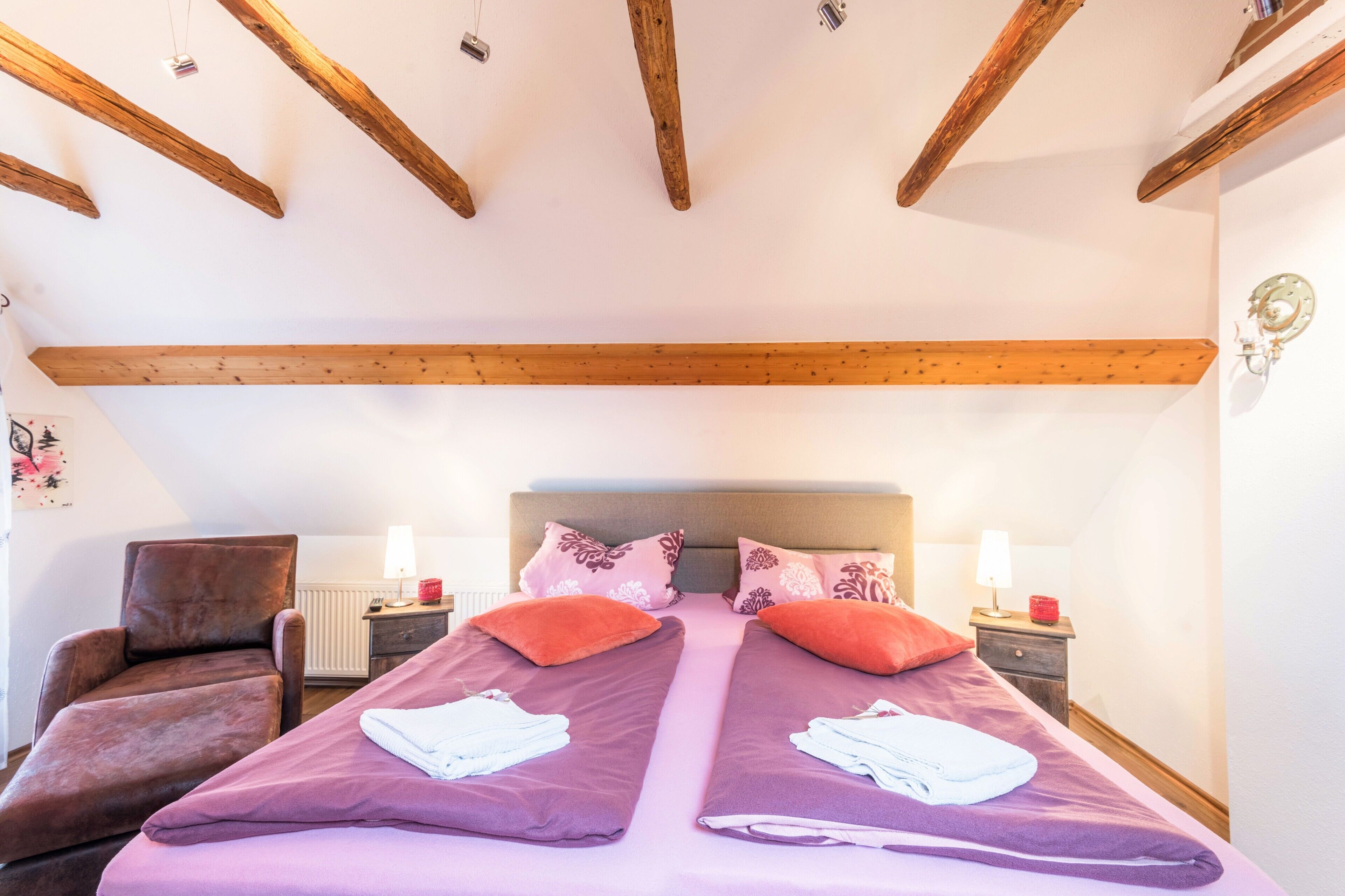Ein Doppelbett mit zwei Kissen, ein Sessel, Nachttische mit Lampen und Holzbalken an der Decke.
