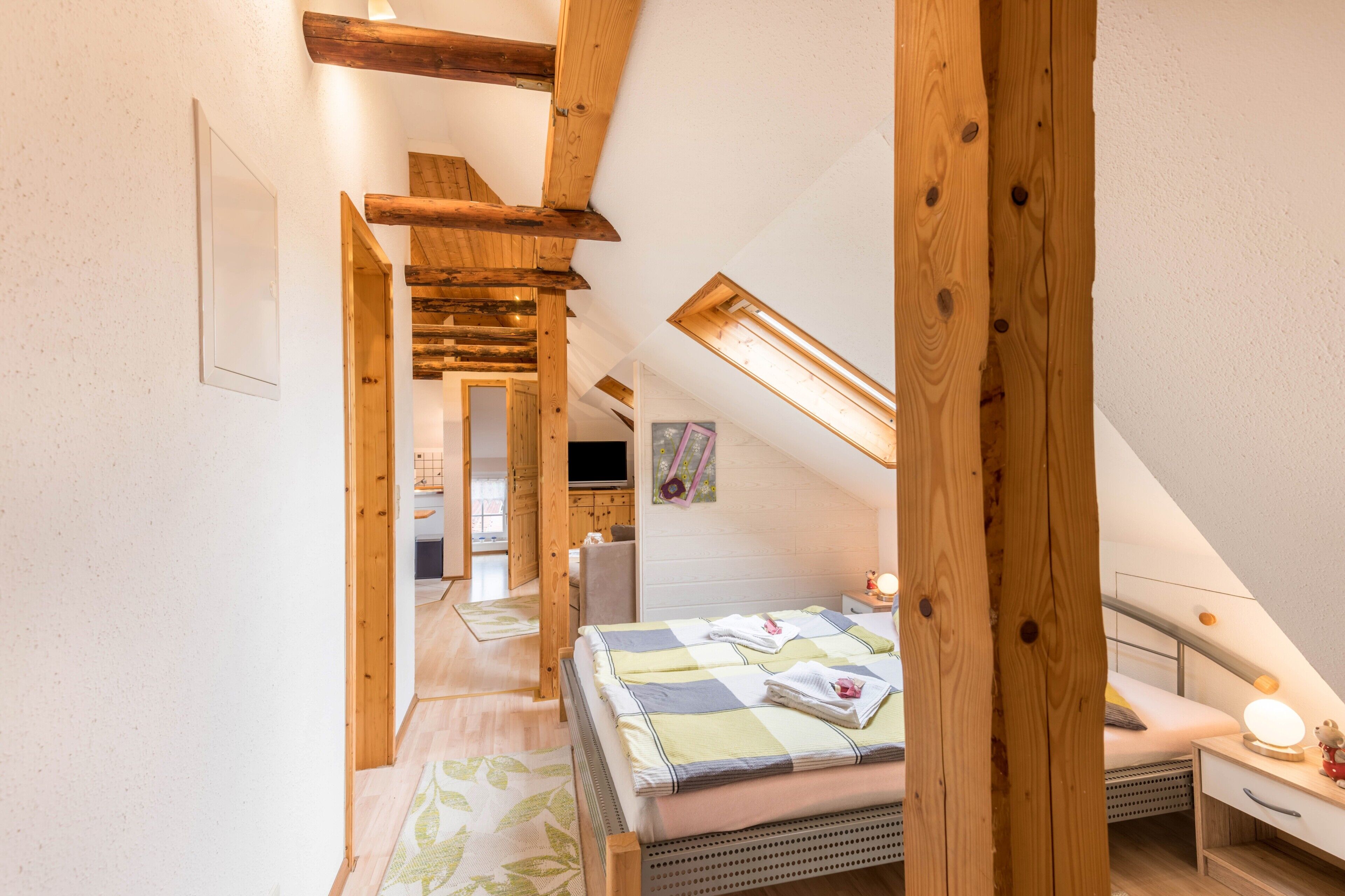 A bedroom with a sloped ceiling, wooden beams, a bed, a desk, and a small table with a lamp.