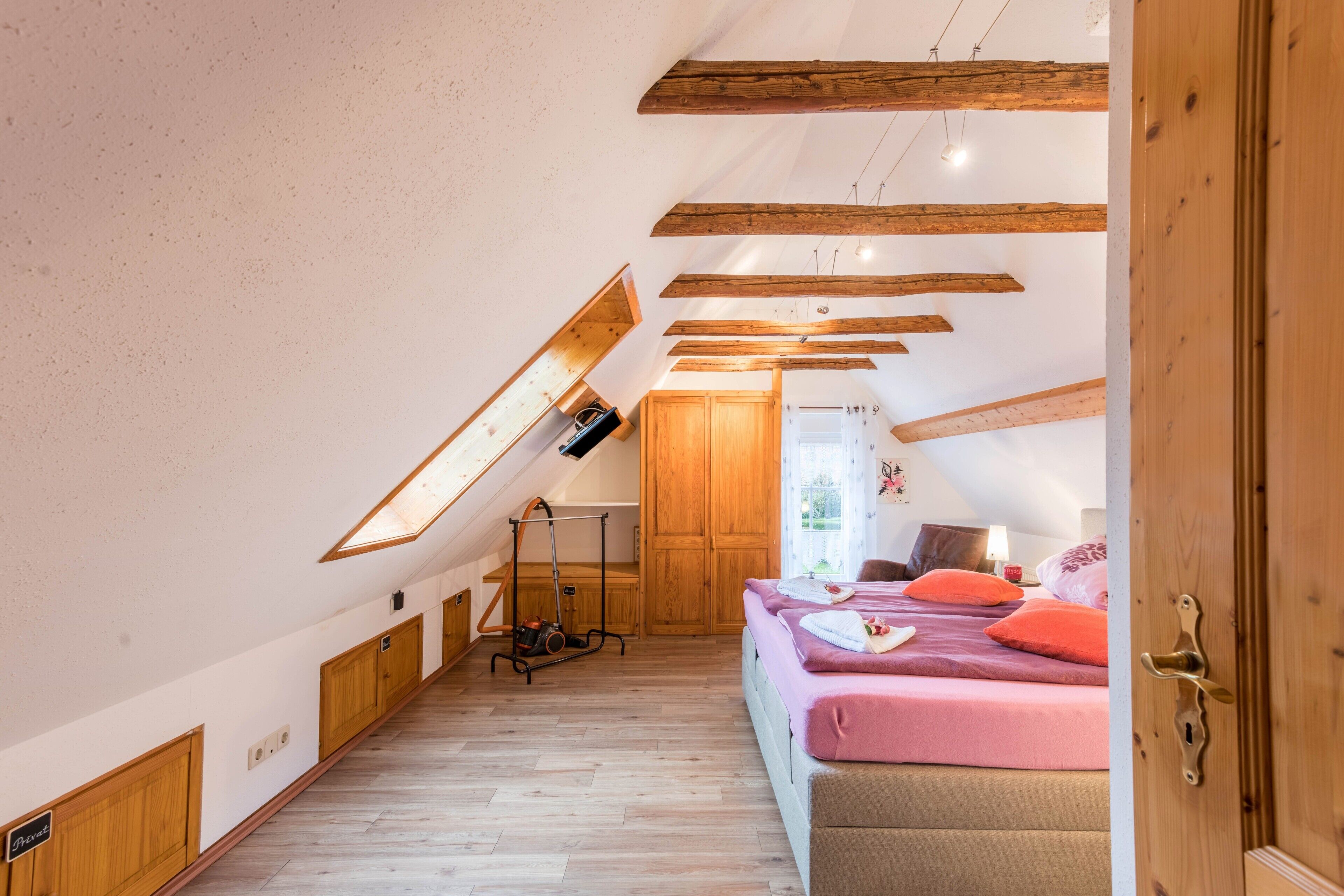 Une chambre avec un plafond en pente, des poutres en bois, un lit avec une literie rose et une armoire en bois.