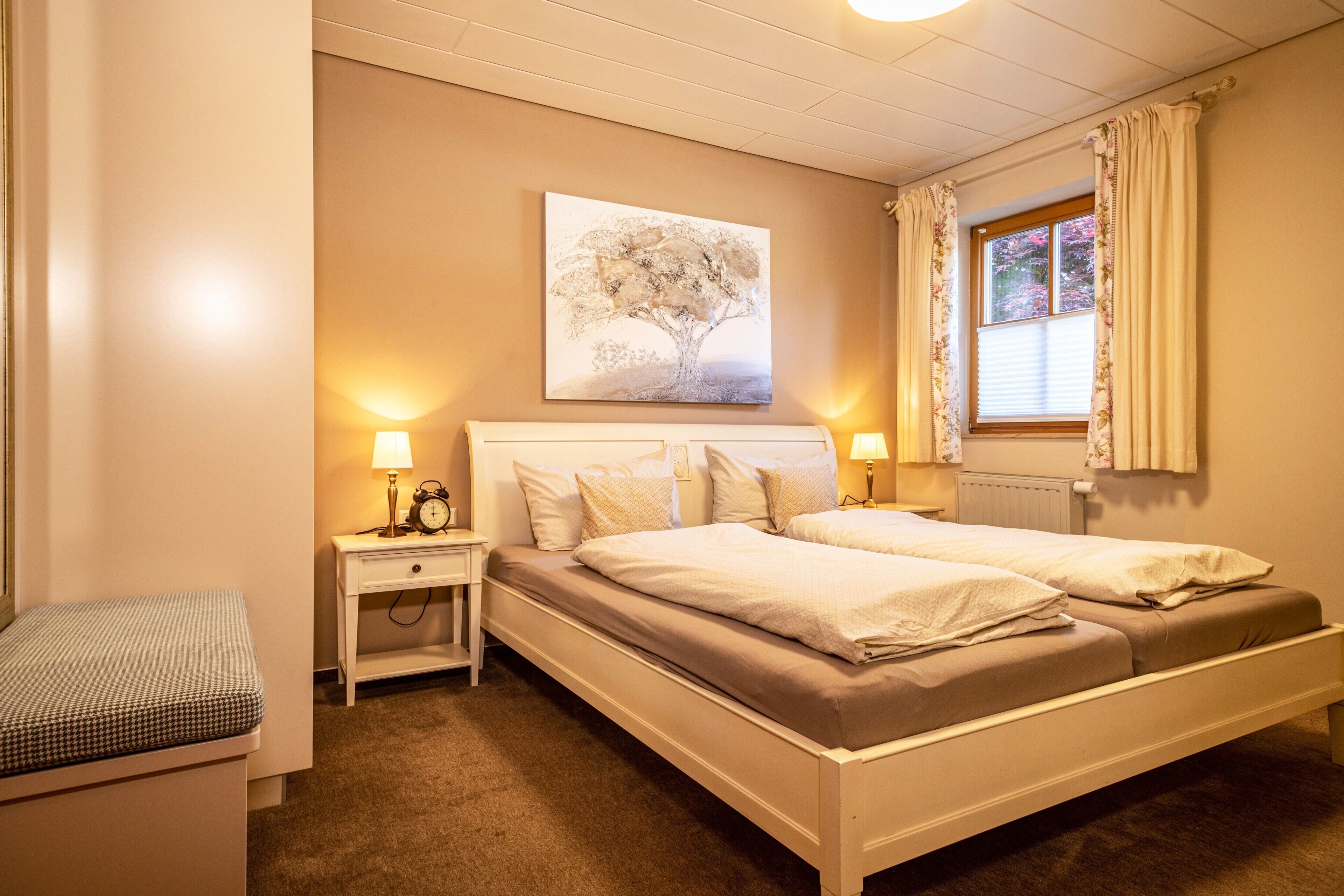 A bedroom with two beds, bedside lamps, a nightstand, a wall-mounted painting, and a window with curtains.