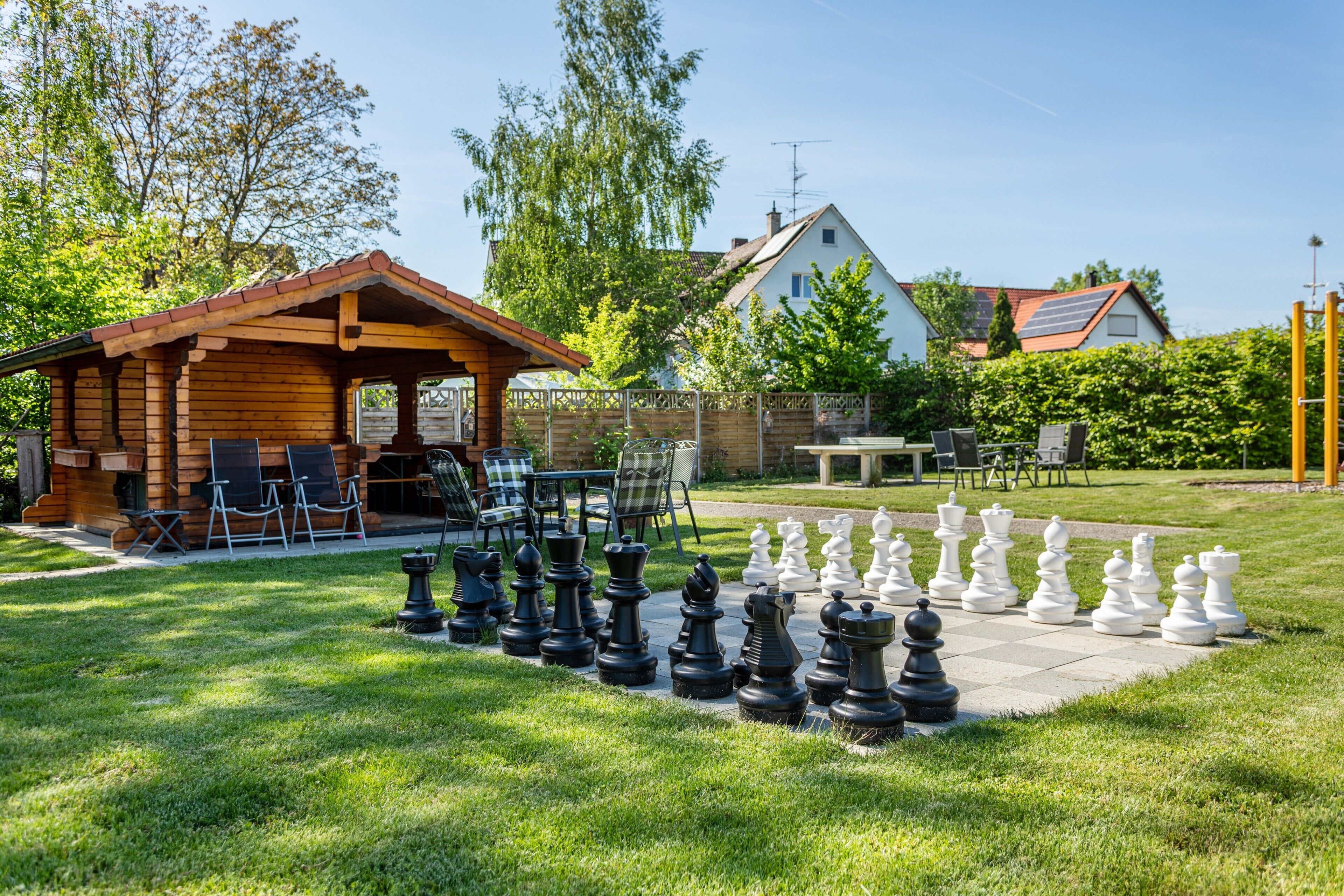 A wooden gazebo with outdoor seating, a chessboard, and a house in the background.