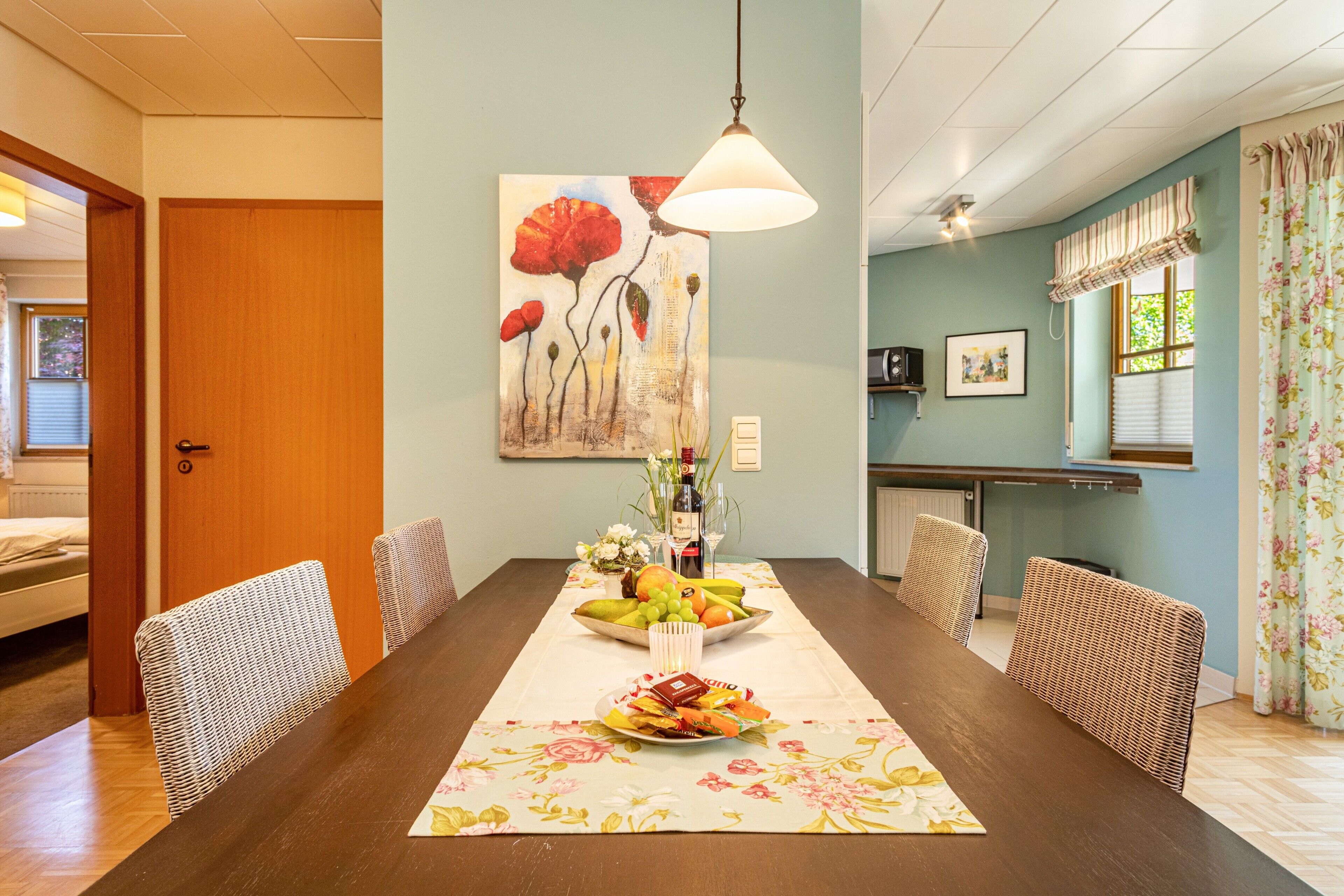 Ein Essbereich mit Holztisch, Korbstühlen, einer Tischläufer mit Blumenmuster und einem Bild an der Wand.