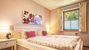 Un dormitorio con una cama doble, lámparas de noche, mesitas de noche, un cuadro floral y una ventana con cortinas.