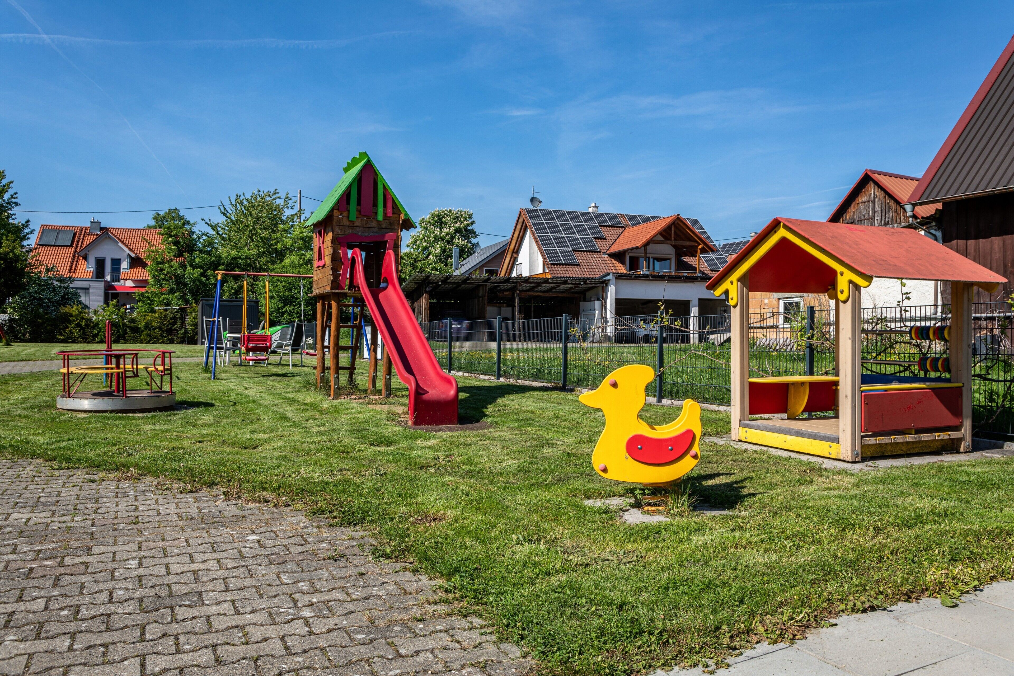Un parc infantil amb tobogan i cadiretes, una estructura de joc amb forma de pato i un petit refugi semblant a una casa.