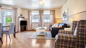 Una sala de estar con televisor, sofá y sillones, un comedor con mesa y sillas, y un ventanal con cortinas.
