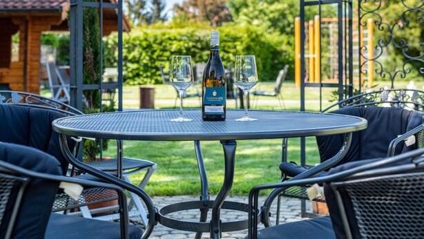 Mesa redonda metálica con dos copas de vino y una botella, rodeada de sillas con cojines oscuros. El conjunto está al aire libre, con vistas a un jardín y un edificio al fondo.