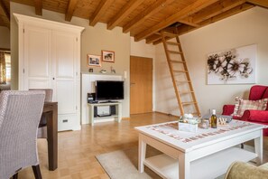 Un salon avec un plafond en bois, un téléviseur posé sur un meuble blanc, un canapé rouge et une table blanche agrémentée d’objets décoratifs.