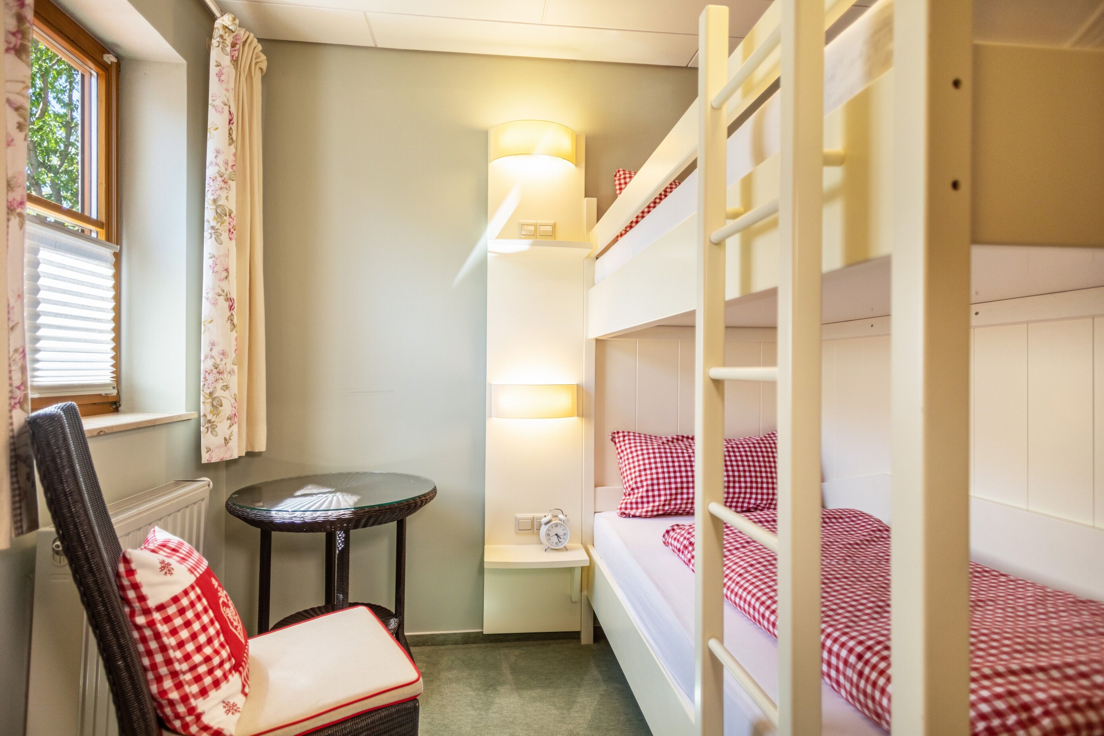 A small, cozy room with bunk beds, a small table, a chair, and a window with curtains.