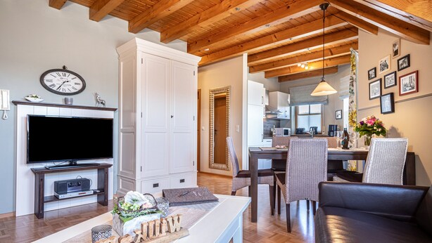 Una sala de estar moderna con televisor de pantalla plana, vigas de madera, un comedor con sillas y una cocina al fondo.