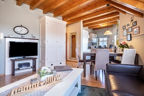 A modern living room with a flat-screen TV, wooden beams, a dining area with chairs, and a kitchen in the background.