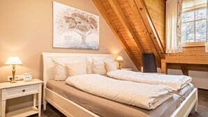 Un dormitorio con techo de madera, cama con ropa de cama blanca, mesita de noche con lámpara y un cuadro de un árbol.