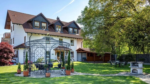 Una casa de dos pisos con un porche cubierto, un jardín con plantas en macetas y una pérgola.