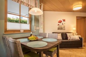 Un coin repas avec une table en bois, des chaises, une fenêtre avec des stores, un canapé et un tableau accroché au mur.