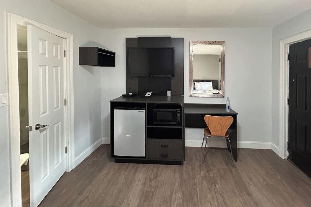 A compact room with a kitchenette, a small desk, a chair, a mirror, and a door.