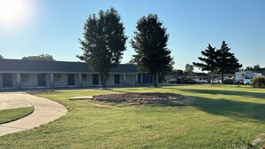 A well-maintained grassy area with a circular flower bed, a paved walkway, and a row of trees. - Motel 6 Ponca City, OK (Ponca City)