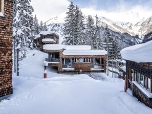 A modern chalet with a brick exterior and multiple balconies, surrounded by snow-covered trees and mountains. - 4-star apartment directly at the piste (Kals am Großglockner)
