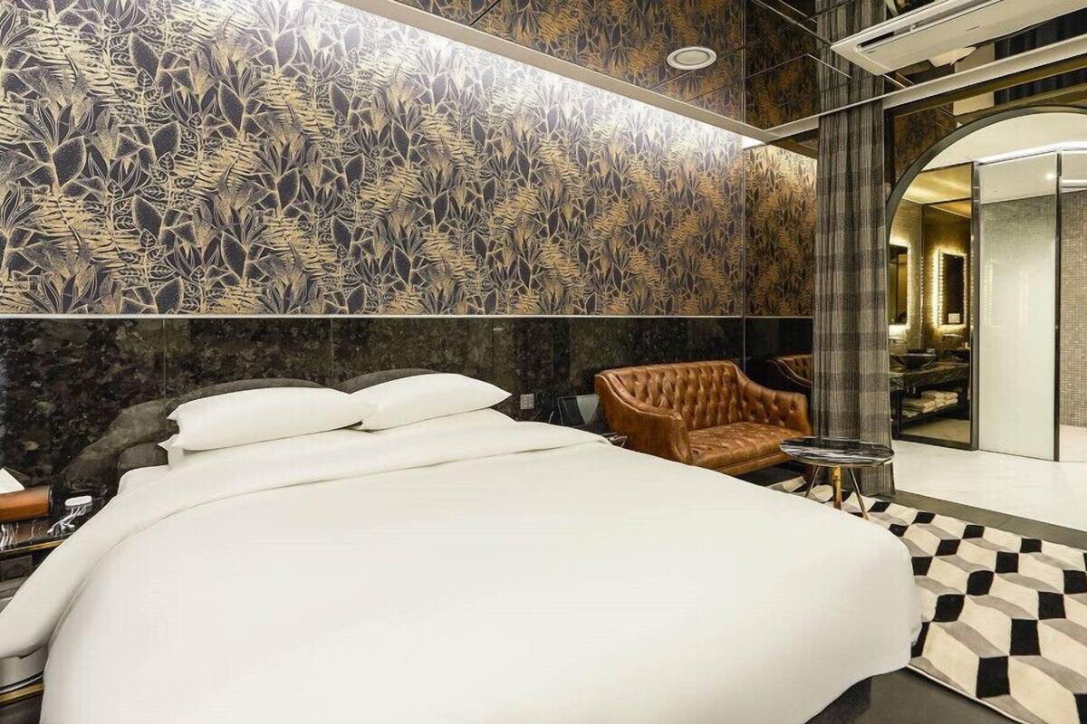 A modern hotel room with a large bed, a seating area with a brown chair, a bathroom with a mirror, and a patterned wall.
