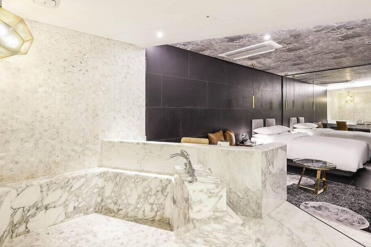 A modern bathroom with a marble tub, a large mirror, and a separate bedroom area with two beds.
