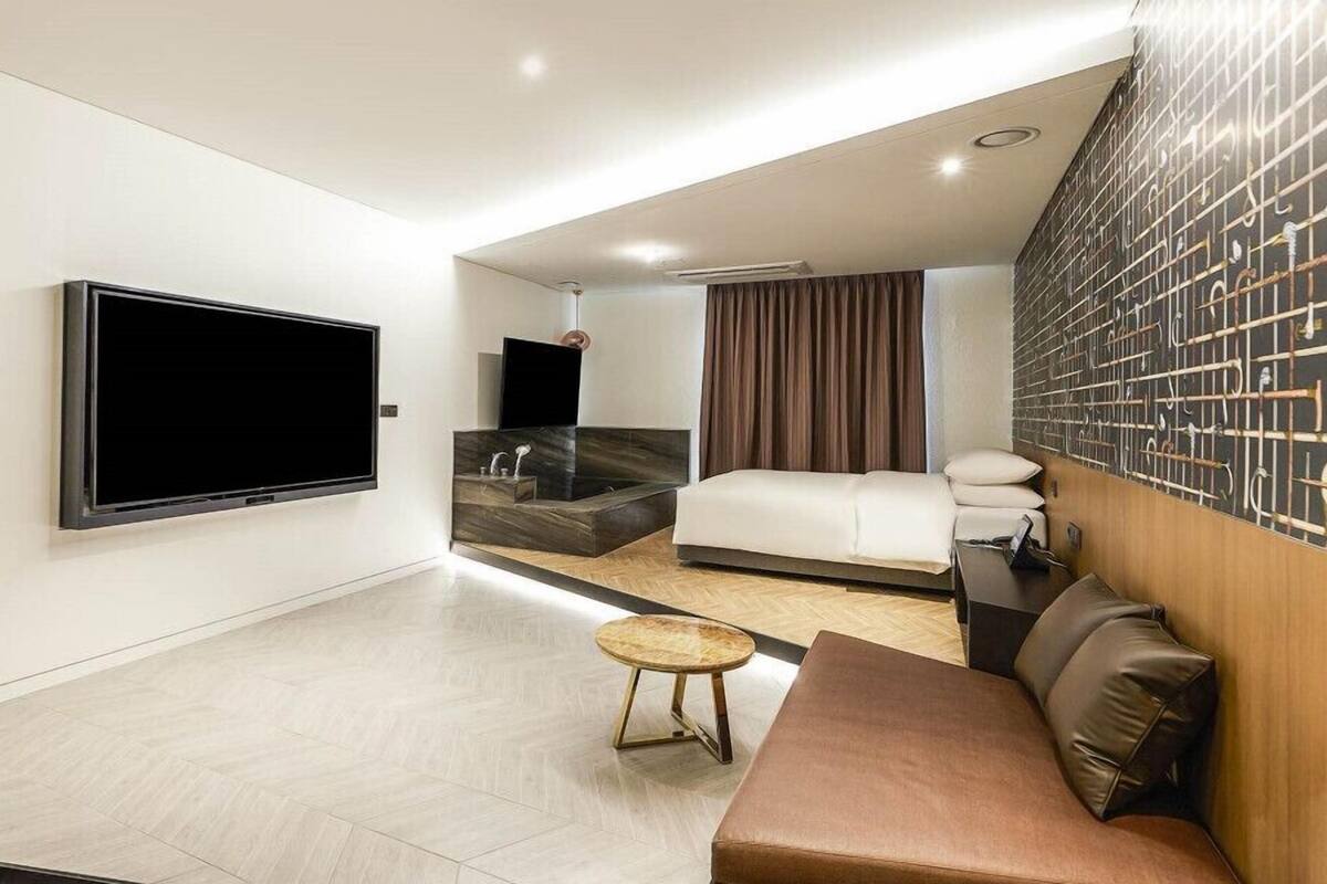 A modern hotel room with a large flat-screen TV, a bed, a sofa, a small round table, and a wall-mounted towel rack.