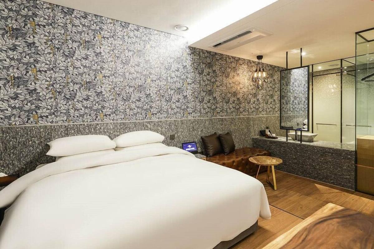 A modern hotel room with a large bed, a small seating area, a glass shower enclosure, and a marble-patterned wall.