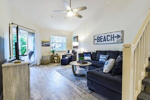 Una sala de estar amplia con un sofá de color oscuro, una mesa de centro y un ventilador de techo. Hay un cartel en la pared que dice 'BEACH' con una flecha señalando a la derecha.