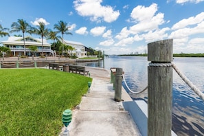A waterfront walkway with a grassy area, wooden posts, and a view of a large building with multiple balconies and palm trees in the background. - Siesta Bayside 3BR Villa: Rare, Spacious Layout, Pickleball, Heated Pools, & Private Beach Access (Siesta Key)