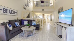 A spacious living room with a grey sofa, a coffee table, and a TV displaying a beach scene. A staircase leads to a balcony with a railing. - Siesta Bayside 3BR Villa: Rare, Spacious Layout, Pickleball, Heated Pools, & Private Beach Access (Siesta Key)