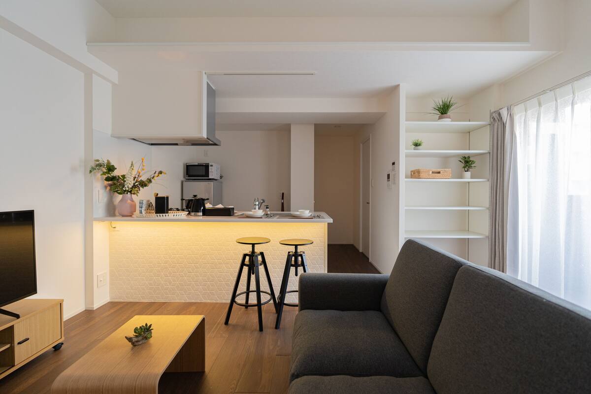 A modern living room with a grey sofa, wooden coffee table, and a TV on the left. A kitchen with a white countertop and two stools is visible in the background.