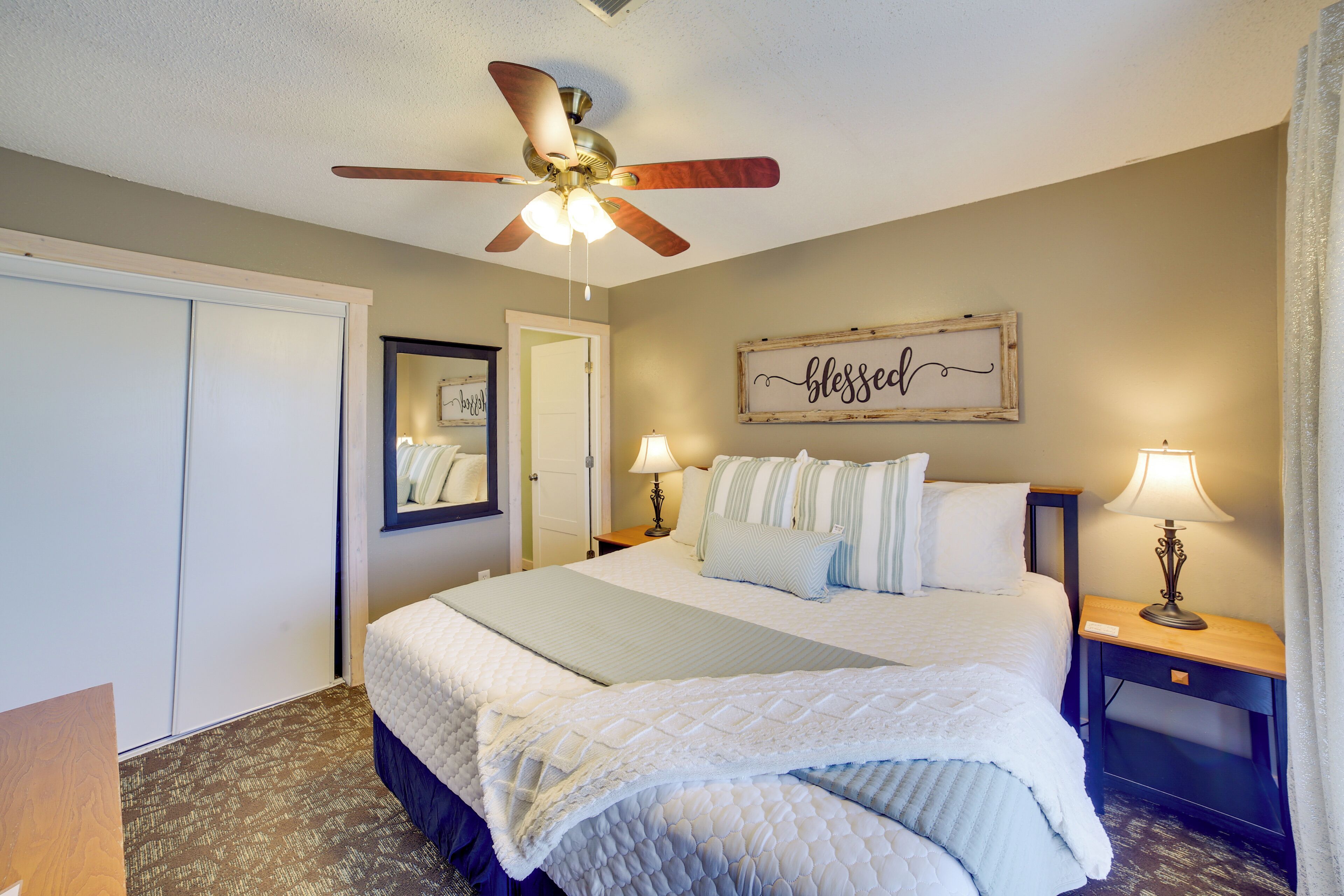 A bedroom with a bed, a ceiling fan, a bedside table with a lamp, a door, and a wall decoration.