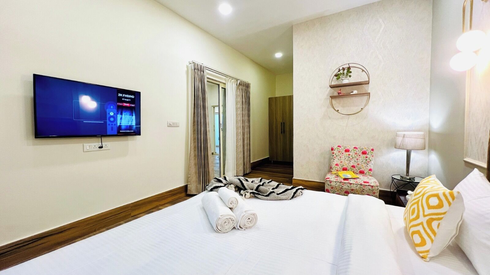A hotel room with a bed, a TV mounted on the wall, a small table with a lamp, a chair, and a wall-mounted shelf with decorative items.