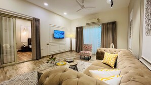 A well-lit living room with a sofa, a coffee table, and a TV. There is a ceiling fan and an air conditioning unit.