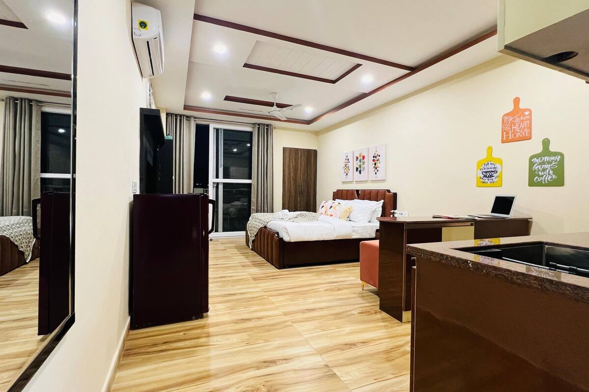 Hotellihuoneessa on sänky, työpöytä ja pieni keittiönurkkaus. Siellä on myös verhot, kattotuuletin ja seinäkoristeita.