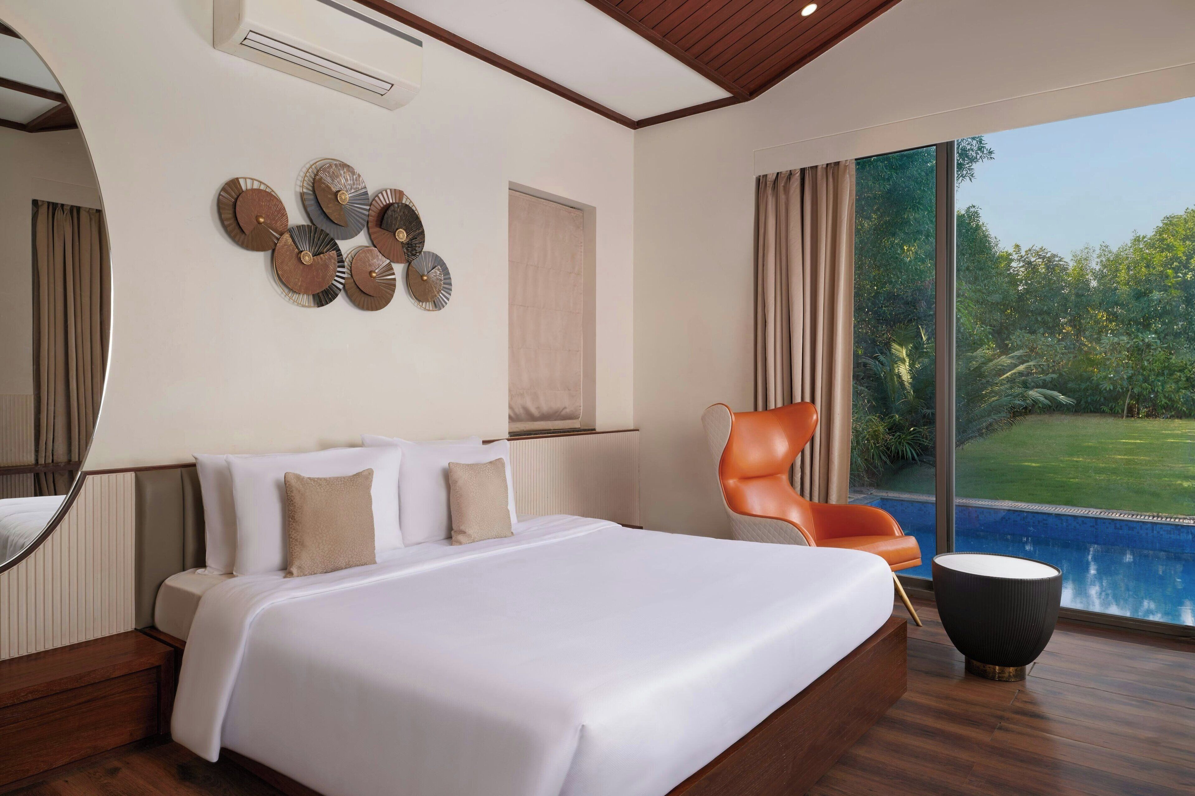 A modern hotel room with a large bed, a seating area with a chair, a large window with a view of a pool and greenery, and decorative wall art.