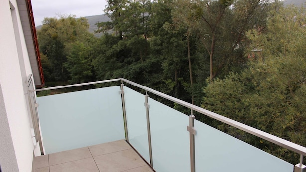 Un balcon doté d’une barrière en verre et d’un sol carrelé, offrant une vue sur une forêt dense et, au loin, sur une montagne.