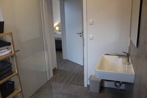 Een badkamer met een wit wastafelmeubel, een kleine prullenbak en een houten plank met opgevouwen handdoeken.