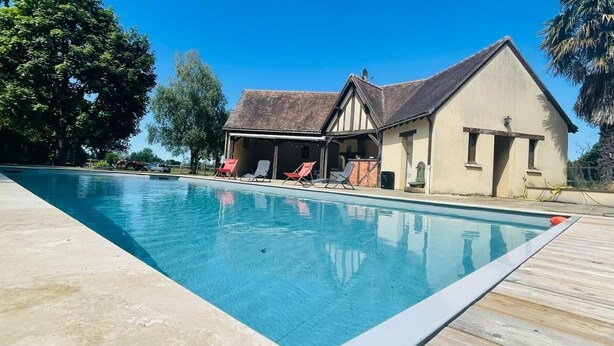 Une piscine avec des chaises longues, un espace de détente couvert et un bâtiment doté d’un toit en pente.
