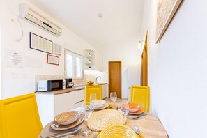 Une cuisine compacte avec des meubles blancs, un four à micro-ondes et une machine à café. On y trouve une table à manger avec des chaises jaunes et un set de table en fibres tressées.