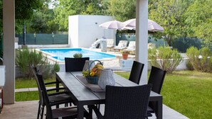 En udendørs terrasse med spisebord og stole, en swimmingpool og liggestole i baggrunden.