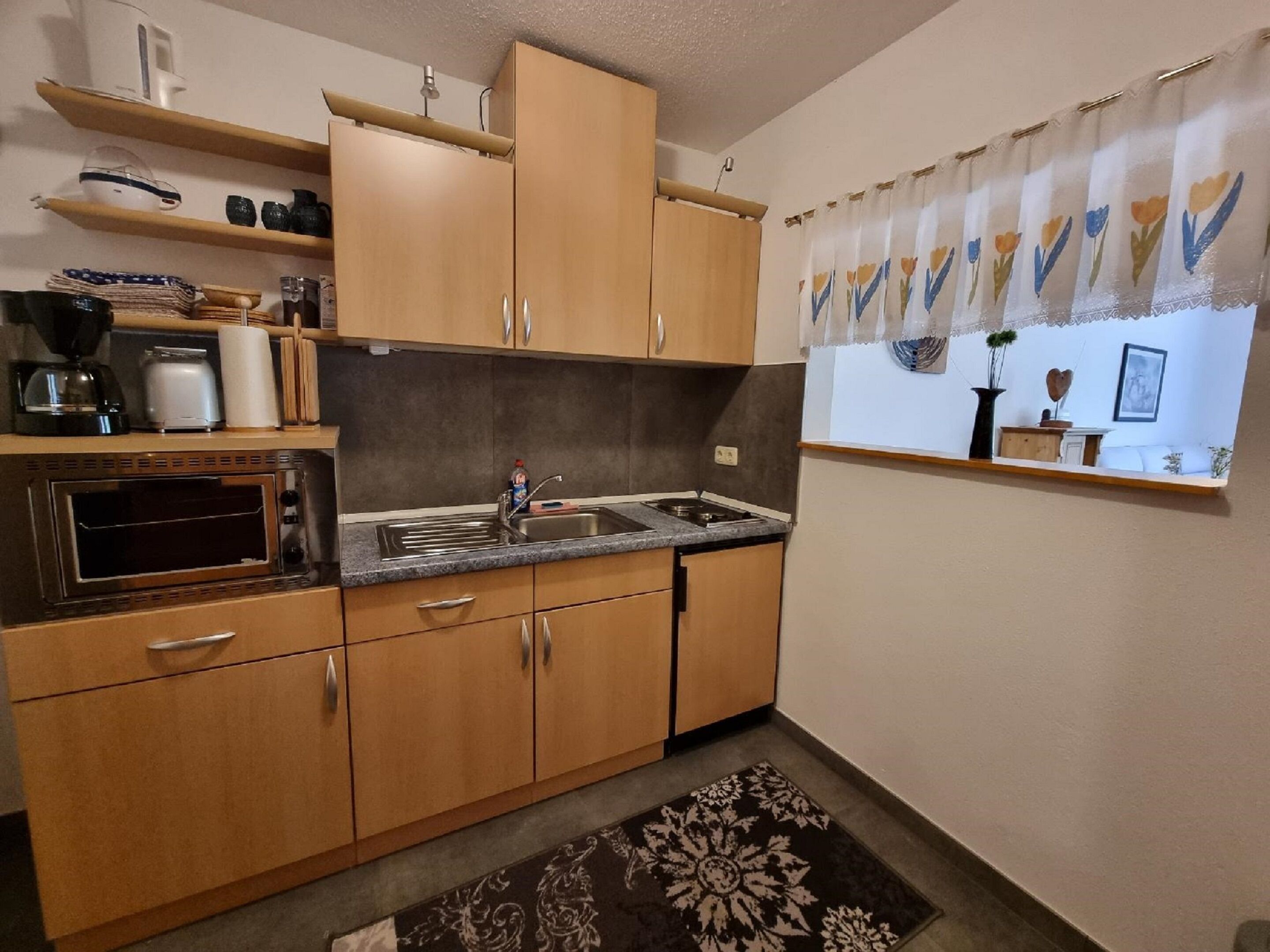 Una cucina compatta con mobili in legno, forno integrato, lavandino e finestra con tenda a fiori.