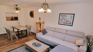 A modern living room with a sofa, coffee table, dining table, chairs, and a chandelier. - Cafe Altmühl, Ferienwohnungen Gästehaus Krimhilde direkt am Wasser (Essing)