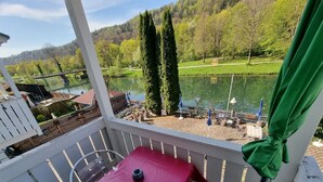 A balcony with a red table, a chair, and a view of a lake and forest. - Cafe Altmühl, Ferienwohnungen Gästehaus Krimhilde direkt am Wasser (Essing)