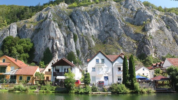 A lakeside village with traditional buildings, a café, and a castle on a hill. - Cafe Altmühl, Ferienwohnungen Gästehaus Krimhilde direkt am Wasser (Essing)