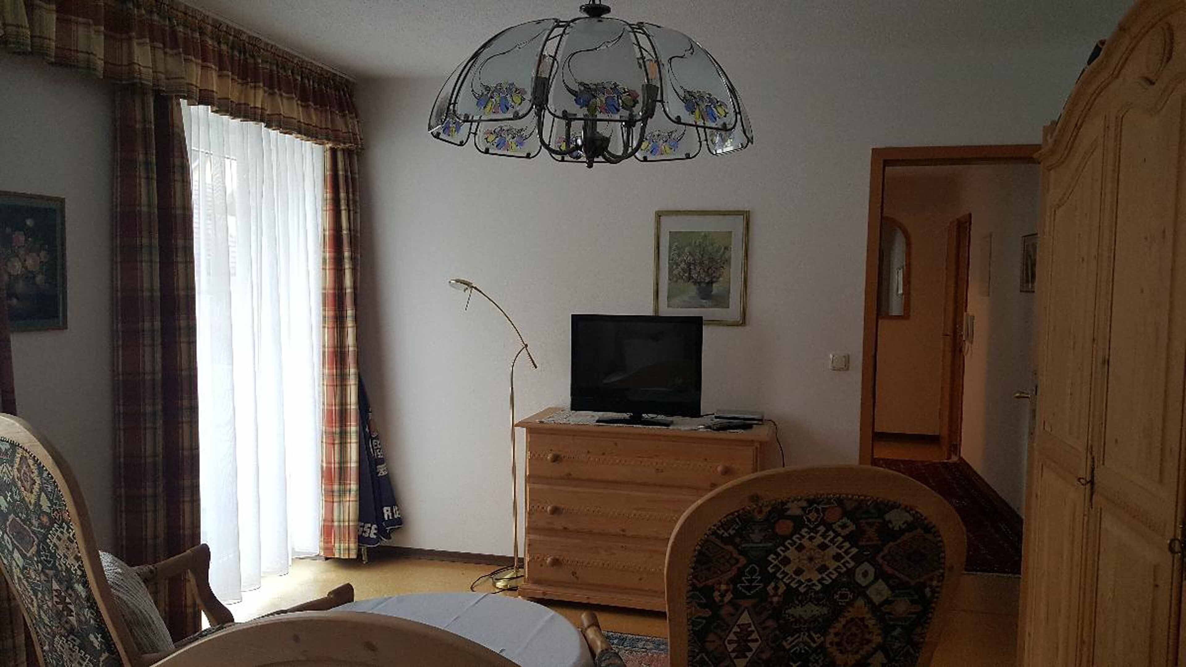 A room with a wooden dresser, a television on top, a patterned chair, a chandelier, and a door leading to another room.