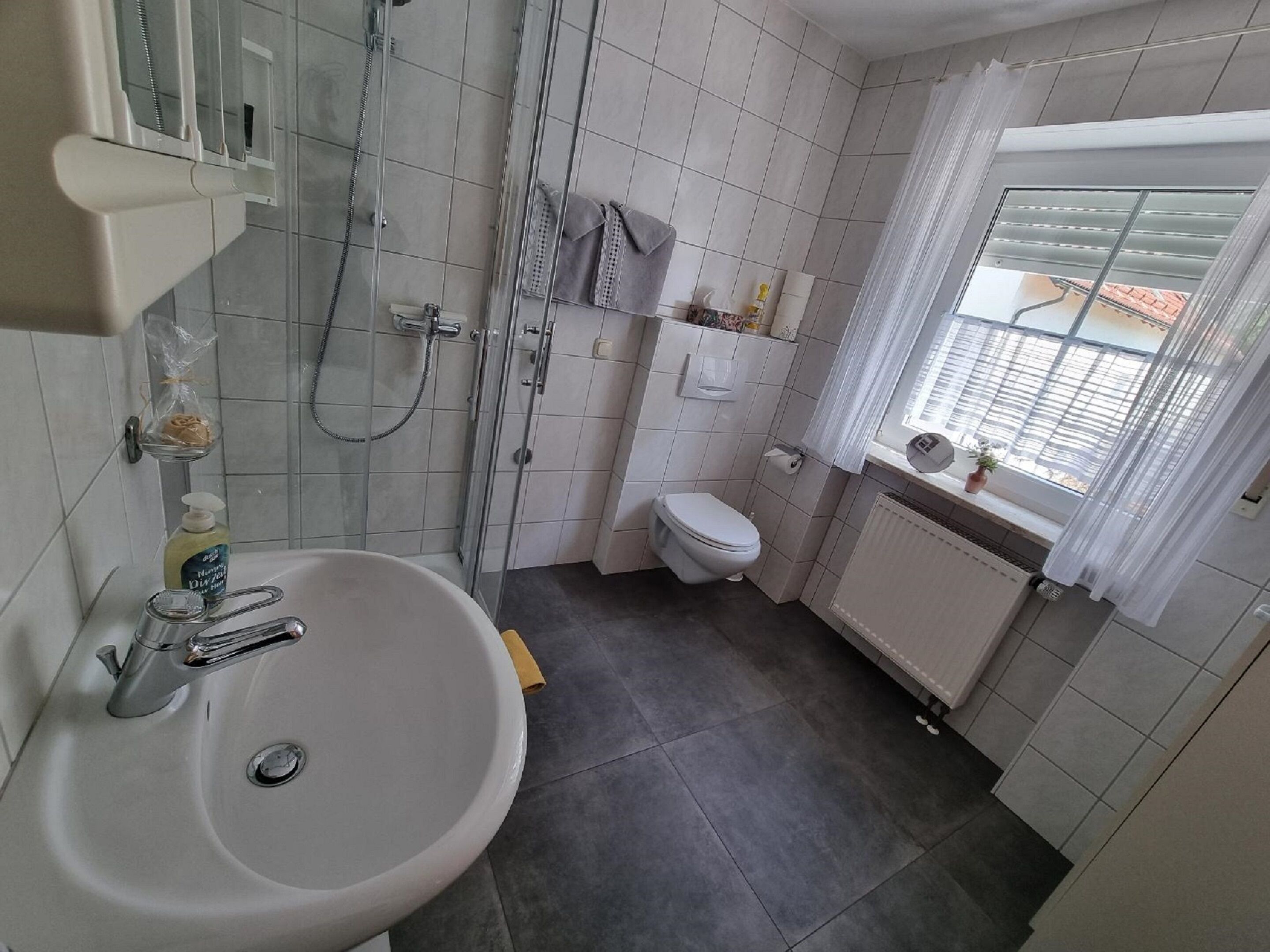 A bathroom with a white sink, a glass shower enclosure, a toilet, and a window with a view of buildings outside.