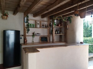 A rustic bar area with a black refrigerator, shelves with bottles, a chalkboard menu, and hanging baskets.